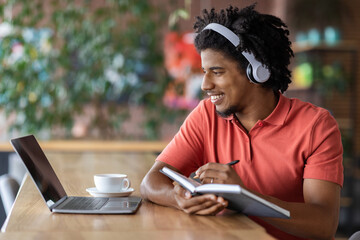 Smiling young black curly man in wireless headphones makes notes in notebook and looks at laptop in cafe interior. Work remotely for digital nomad, modern education and learn language, online lesson