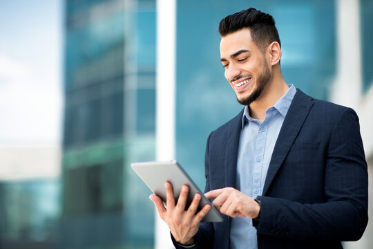 Successful arab guy in formal wear standing by office building, holding digital tablet and smiling, looking for new business opportunities online, using business application, copy space, panorama