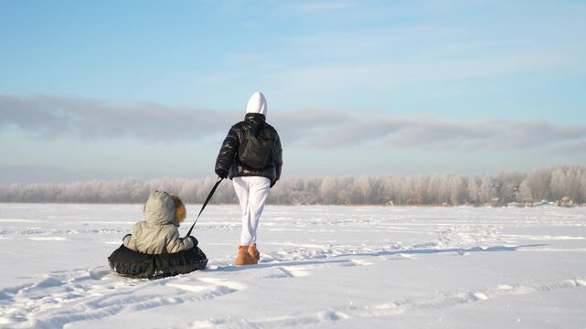 Mom drags her son on a tubing sled. Sunny winter weather.