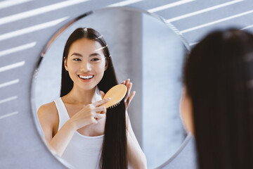 A woman is brushing her long, straight hair while looking at herself in the mirror. Sunlight streams through the window highlighting her features. It is morning in a modern bathroom.