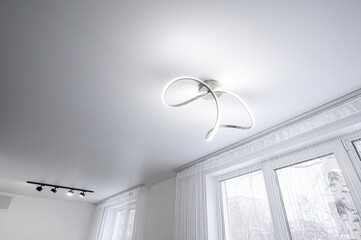 modern room with a sleek white ceiling light fixture featuring curved LED design, complemented by track lighting. Large windows with sheer white curtains let in natural light