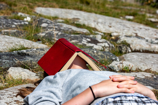 Anonymous bored girl asleep on rocks with a book over her face.