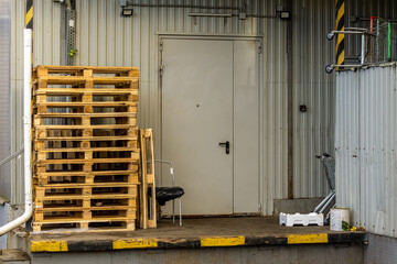 Stack of wooden pallets stands on loading dock in front of metal door. Shopping cart and industrial wall elements visible nearby.