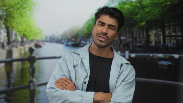 Man stands with crossed arms on street beside canal, leaning on metal railing under leafy trees with closed eyes; confidence.