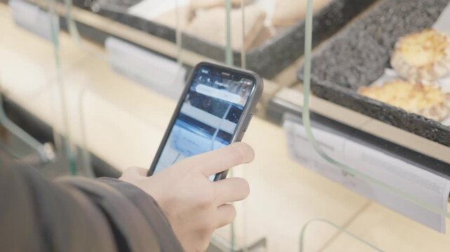 A close-up shot of a hand holding a mobile phone, browsing dessert options displayed behind glass. The modern setting captures the intersection of technology and food choices in a vibrant environment.