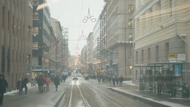 A bustling urban street during winter, adorned with festive holiday lights. Snow covers the ground as people walk along, creating a vibrant and warm atmosphere despite the chilly weather.