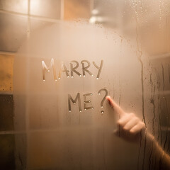 Fototapeta premium Woman hand writing marry me question on wet steam mirror surface. Romantic marriage proposal concept for valentine day. Love confession text message on fogged glass with blurred warm light background