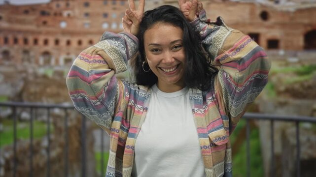 Woman smiling with tongue out showing peace sign with both hands behind head at roman ruins building, wearing patterned jacket and white tee; joy travel memories.