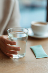 Woman's hand holding a glass of water next to a coffee cup and a supplement packet
