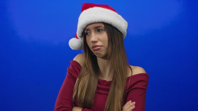Young hispanic woman wearing santa hat crosses arms with pouting lips in blue studio; disappointment.