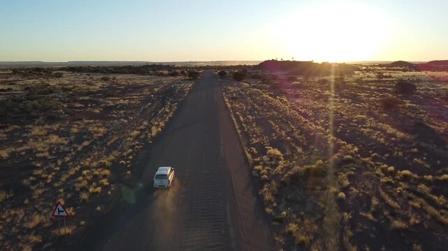 Lone white vehicle traveling across the rocky desert of Namibia leaving a cloud of dust on a gravel road; aerial drone perspective of transport in a remote African wilderness during sunset