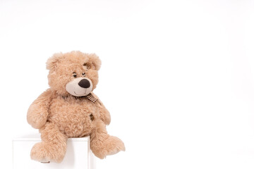 Fototapeta premium Cute brown teddy bear sitting on a white cube against a clean white background with generous copy space. The soft plush texture and warm neutral tones create a cozy, comforting childhood atmosphere