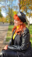 A beautiful red-haired cheerful girl in a black coat and hat sits on a bench in an autumn park