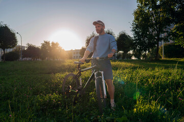 Guy with sunglasses stands with his bicycle in a grassy field at sunset. He is wearing casual clothes and has a backpack. The scene shows a park landscape during golden hour