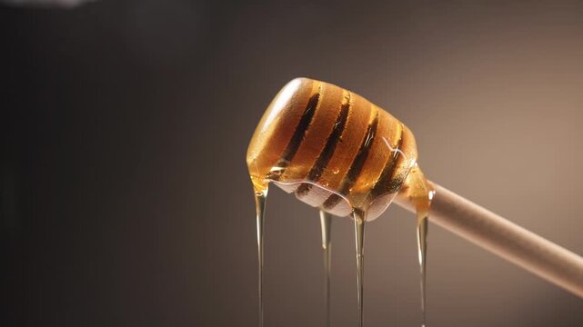 Slow motion slo mo of raw honey bee syrup from beehive with glowing yellow texture falling on top of honeycomb comb with wooden stick and droplets fall slowly for food preparation of sweet treat