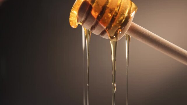 Slow motion slo mo of raw honey bee syrup from beehive with glowing yellow texture falling on top of honeycomb comb with wooden stick and droplets fall slowly for food preparation of sweet treat