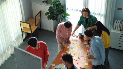 Top view of diverse business people writing and stick sticky notes at whiteboard. Creative team working together and brainstorming marketing idea for startup project at table with notes. Convocation.