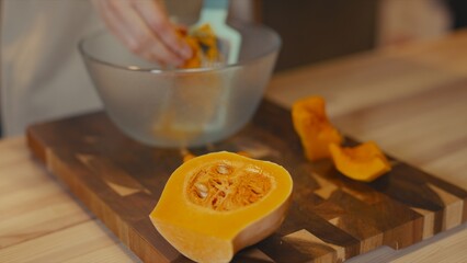 Close-up of butternut squash slices on a wooden cutting board. In the blurred background, a woman grates fresh organic squash into glass bowl. Seasonal healthy food preparation on rustic wooden table.