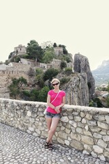 Fototapeta premium Female tourist posing for a photo on a stone wall overlooking the historic Guadalest castle ruins and mountain village architecture in Spain on a sunny day