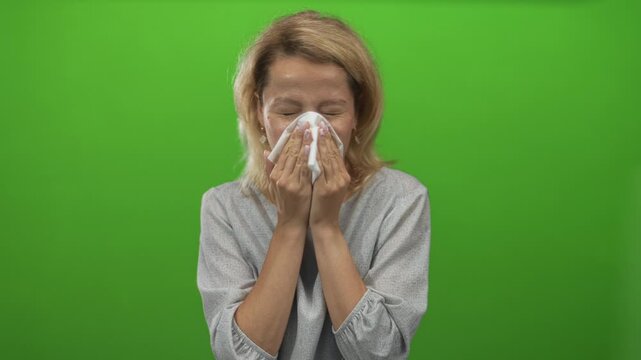 Young blonde woman blowing her nose into a tissue on bright green studio set; discomfort.