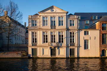 Elegant neoclassical canal house in Bruges with Roman numeral pediment and tall white windows reflecting warm golden hour light on the water