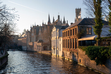 Golden hour view of Bruges canal with medieval brick buildings and gothic spires reflecting in rippling water, historic old town cityscape in Belgium