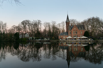 Lakeside neo-gothic mansion in Bruges reflected in calm water at blue hour with bare winter trees and soft pastel evening sky