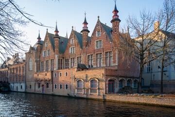 Historic gothic brick building on Bruges canal in warm evening light, medieval European architecture reflected in calm river water, Belgium old town cityscape