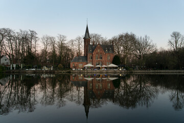 Romantic neo-gothic lakeside castle restaurant in Bruges reflected in calm water at twilight with bare winter trees and pastel evening sky