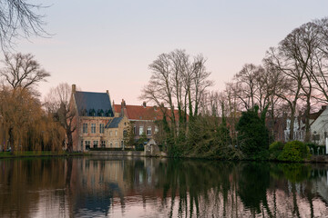 Tranquil Bruges lakeside houses and bare winter trees reflected in calm water at pastel dusk, historic Belgian architecture by Minnewater Lake