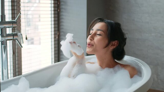 A woman sits in a bathtub filled with bubbles. She smiles while holding a bubble in a bright bathroom with a window letting in sunlight. The setting is clean and stylish.