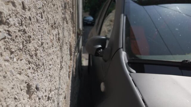 Black car parked extremely close to an old wall. Experienced driver shows precise parking on a narrow Mediterranean street. Tilt up shot reveals the tiny space between vehicle and wall.