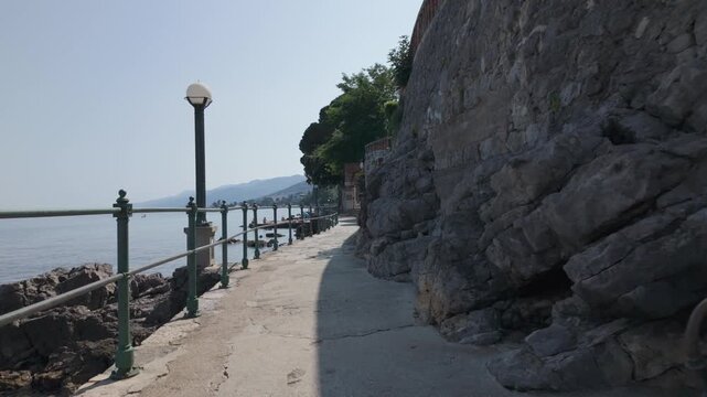 Walking along the Lungomare Coastal Promenade in Volosko, Croatia on a sunny summer day. Rocky Adriatic Sea coastline with railings and blue water. The famous seaside walkway is over 100 years old.