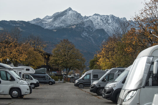 Reihe geparkter Wohnmobile und Reisemobile auf einem asphaltierten Stellplatz, beidseitig aufgereiht; im Hintergrund hohe, teils schneebedeckte Berggipfel. Herbstliche B&auml;ume mit farbigem Laub rahmen d