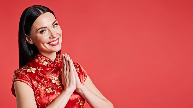 Smiling woman in traditional red cheongsam dress with golden floral patterns, hands together in greeting or prayer gesture