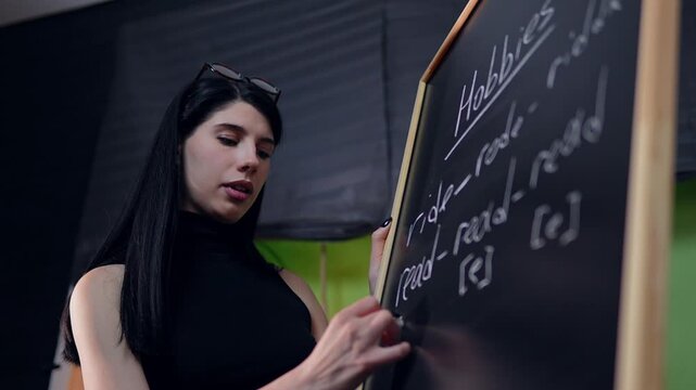 Closeup view of pretty Caucasian woman writing irregular English verbs on the blackboard filmed in slow motion. Female teacher is explaining grammar standing in a room and making notes with a chalk