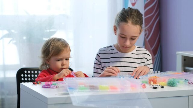 Two sisters are at home making colorful bracelets with rubber bands. The older sister helps her younger sibling choose colors and create fun designs. They share a special moment together