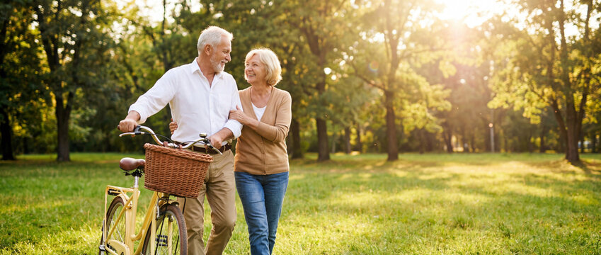 Happy senior couple walking with bicycle in sunny park during golden hour