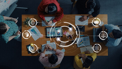 A top-down view of a diverse team engaged in collaborative work, using laptops and discussing innovative ideas with a digital interface overlay showcasing business strategies. Trope