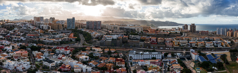 Naklejka premium Aerial view shows Las Palmas de Gran Canaria with layered neighborhoods, tall towers by the waterfront, curving roads, terraced hills, and harbor under late afternoon clouds.
