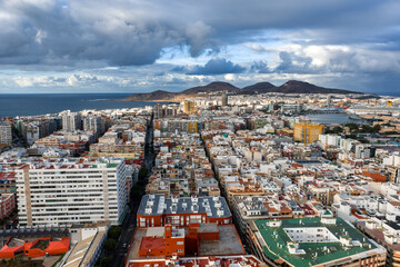 Obraz premium Aerial cityscape of Las Palmas de Gran Canaria shows white and pastel blocks, La Isleta twin peaks, port basin, cruise ships, winter light, and dramatic clouds.