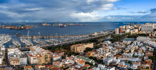 Aerial view shows Muelle Deportivo yachts, cargo cranes, and rigs in Las Palmas de Gran Canaria. Tankers and container ships sit on the Atlantic under partly stormy skies.