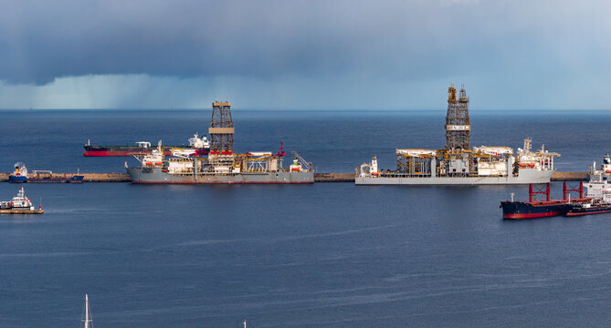 Las Palmas de Gran Canaria harbor shows two offshore drillships at a breakwater, with cargo vessels, tugboats, and tankers waiting on calm water under distant rain.