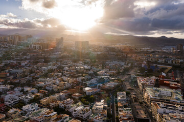 Aerial view of Las Palmas de Gran Canaria at golden hour, sun rays pierce clouds, whitewashed houses, terracotta roofs, gridded streets, towers by waterfront, bay at right. © Aerial Film Studio