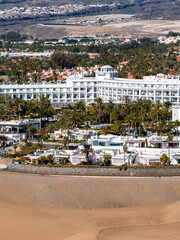 Fototapeta premium Aerial view of Maspalomas Dunes in Gran Canaria with the white RIU hotel, beachfront hotels, villas, pools, stone seawall, palm groves, and distant hills in bright midday light.