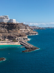 Fototapeta premium High angle view of Amadores Beach near Puerto Rico de Gran Canaria shows rugged cliffs, stepped hotels, a curved breakwater, busy sand, clear sky, and calm Atlantic.
