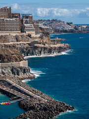 Fototapeta premium Tiered hotels and apartments sit on volcanic cliffs in Gran Canaria near Puerto Rico and Amadores, with a stone breakwater, sun deck, pedal boats, and waves on rock ledges.