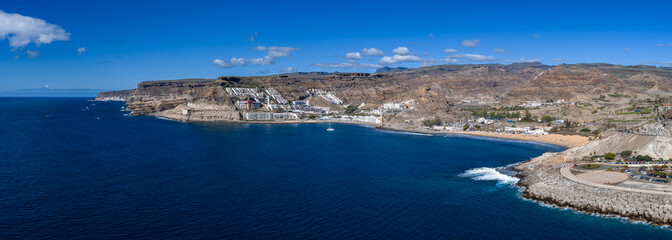Obraz premium Aerial view shows Gran Canaria south coast between Amadores and Puerto Rico. White resorts on arid cliffs, breakwater, roundabout viewpoint, boats, and distant Teide peak visible.