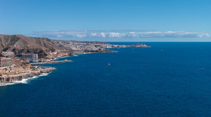 Terraced resorts on volcanic cliffs descend to deep cobalt Atlantic, with marinas, breakwaters, and sailboats near Puerto Rico and Arguineguin under clear midday skies. © Aerial Film Studio