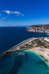 Fototapeta premium Aerial view shows Amadores Beach, Gran Canaria, sunbathers, umbrellas, curving breakwater, marina, inflatable park, boats, terraced hotels, and distant Mount Teide.